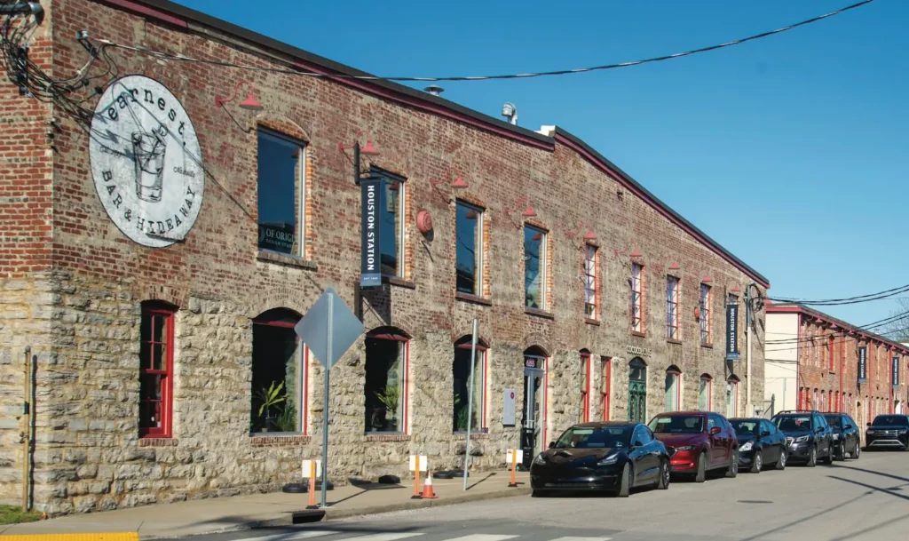 Wedgewood-Houston neighborhood scene featuring brick buildings with industrial architecture, including the "Earnest Bar & Hideaway" sign, vibrant street life, and parked cars along a sunny street.