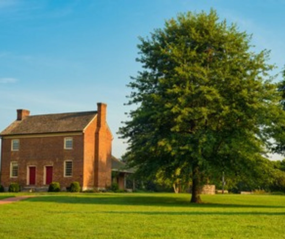 Historic brick house with red doors and large green tree in Goodlettsville, Tennessee, surrounded by open lawn.