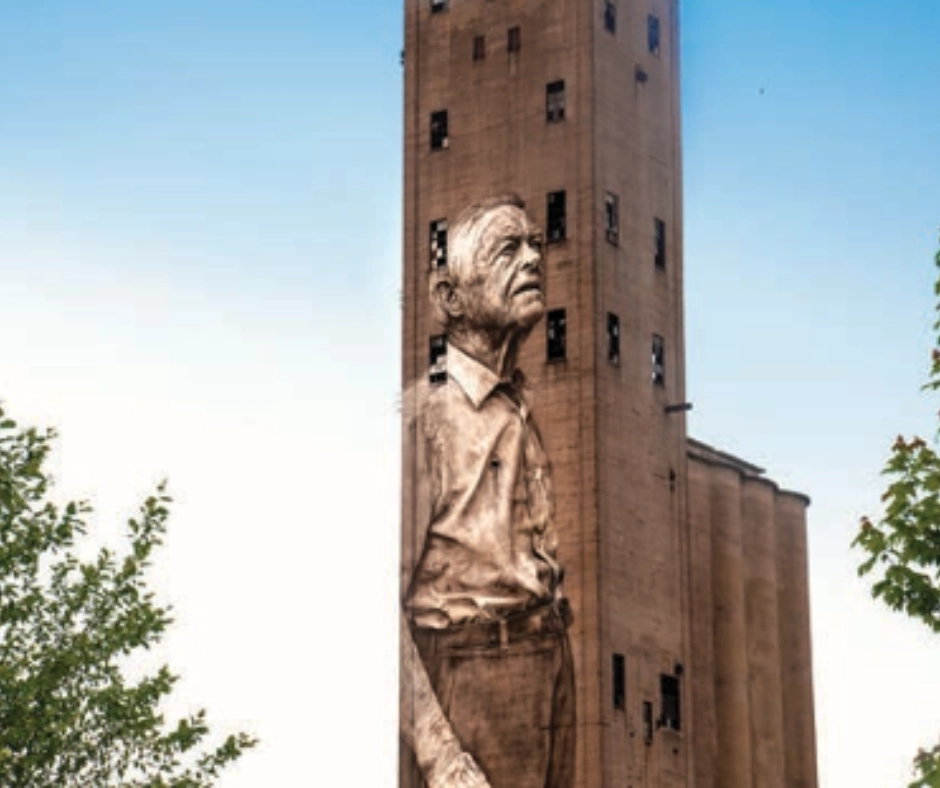 Mural of an elderly man on the side of an industrial building in The Nations neighborhood, Nashville, surrounded by trees and clear blue sky, symbolizing the area's blend of history and modern revitalization.