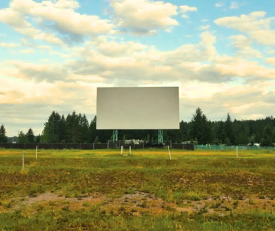 Empty outdoor movie screen in a grassy field surrounded by trees under a partly cloudy sky.